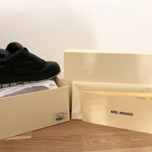 Axel Arigato skor - Säljer ett par svarta puff puff sneakers från Axel Arigato med chunky grå sula och snygga detaljer i guld på sidan. Skorna har snörning, rund tå och är tillverkade i skinn med perforeringar på ovansidan för extra vibe. Perfekt för dig som gillar stilrena och moderna sneakers.