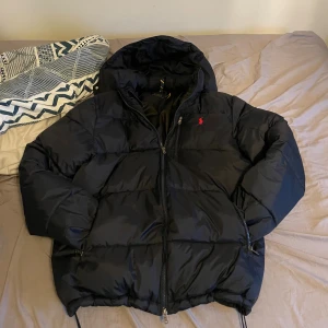 Mörkblå pufferjacka från Polo Ralph Lauren - Riktig snygg mörkblå pufferjacka från Polo Ralph Lauren med huva och dragkedja framtill. Jackan har en röd Polo-logga på bröstet, snörning vid nederkant och två sidofickor med dragkedja. Perfekt för kalla dagar och har en riktigt schysst, puffig look. För mer info eller bilder så är det bara att skriva.