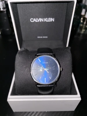 Svart Calvin Klein klocka med läderband - Stilren klocka från Calvin Klein med rund silverfärgad boett och svart urtavla. Klockan har ett svart läderband och minimalistiska detaljer på urtavlan. Perfekt för dig som gillar enkel och modern design.