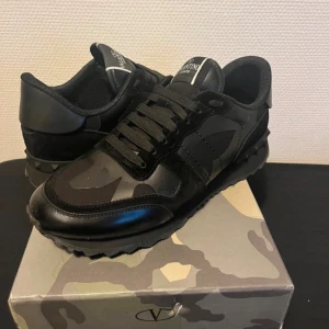 Valentino Garavani Rockrunner sneakers - Snygga svarta Valentino Garavani Rockrunner sneakers med coolt kamouflagemönster. Skorna har detaljer i skinn och mocka, samt svarta snören och en robust sula. Perfekta för dig som vill sticka ut med exklusiva och trendiga sneakers.