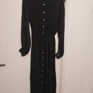 Svart långklänning från The Fated - Svart långklänning från The Fated med knappar hela vägen framtill och lång ärm. Klänningen har ett knytband i midjan med en liten guldfärgad detalj. Tunt, lätt material som faller snyggt och ger en avslappnad men stilren look.