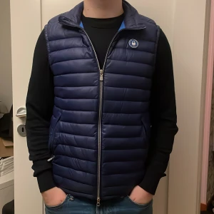 Marinblå dunväst från Sail Racing - Snygg marinblå dunväst från Sail Racing med quiltad design och två dragkedjeförsedda fickor. Västen har hög krage, logomärke på bröstet och hel dragkedja framtill. Perfekt för lager-på-lager och stilren look.