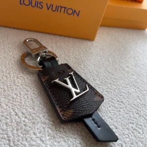  Vuitton brunt skinn nyckelring - Snyggt nyckelring från  Vuitton i mörkbrunt skinn med klassiskt -emblem i silver framtill.  nyckelring har en unik formad läderdetalj och exklusiva metalldetaljer. Perfekt accessoar för att lyfta din outfit med lyxig känsla.