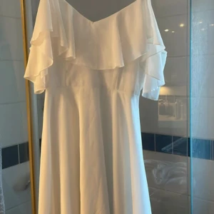 Vit omlottklänning med volang - Superfin vit kortklänning med omlott och volangdetaljer över axlarna. Klänningen har offshoulder-design och är tillverkad i ett lätt, flowy material som ger en luftig känsla. Dragkedja i ryggen för enkel påklädning. Perfekt för sommarens alla tillfällen!