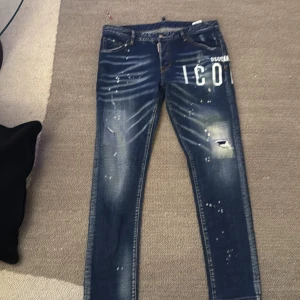 Dsquared2 blå skinny jeans ICON - Snygga blå jeans från Dsquared2 med vit 'ICON'-text på ena benet. Jeansen har skinny passform, slitningar och färgstänk för en cool, trendig look. Klassisk femficksmodell med orange kontrastsömmar och låg midja.