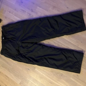 Svarta täckbyxor med dragkedja - Svarta täckbyxor med bred passform och elastisk midja. Byxorna har dragkedja vid bensluten och är tillverkade i ett slitstarkt syntetmaterial som håller dig varm under kalla dagar. Perfekta för vinteraktiviteter och har en enkel, clean look.