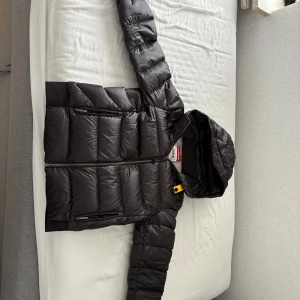 Svart dunjacka från Parajumpers - Säljer en svart dunjacka från Parajumpers, modell Sheen, i storlek Medium. Jackan har huva, dragkedja framtill och två sidofickor med dragkedja. Klassisk pufferstil med ribbade muddar vid ärmslut och detaljerad logotyp på axeln. Y-M