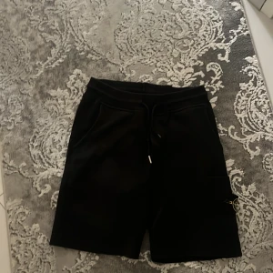 Svarta shorts från Stone Island - Svarta shorts från Stone Island med klassisk logotyp på benfickan. De har snörning i midjan, två sidofickor och en extra ficka med patch. Materialet är mjukt och känns som bomull, perfekt för chill dagar. Snygg och enkel design som funkar till streetwear.