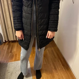 Svart mönstrad pufferjacka Moda Piu Anna - Lång svart pufferjacka från Moda Piu Anna med snygga broderade detaljer och dragkedja framtill. Jackan har ett unikt mönster över hela ytan och quiltad design som ger en cool vibe. Perfekt för dig som vill sticka ut i vinterkylan.