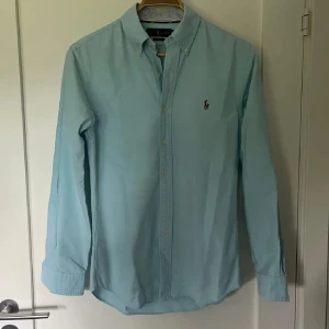 Ralph Lauren skjorta  - Klassisk turkos skjorta från Ralph Lauren med button down-krage och broderad logga på bröstet. Skjortan är i bomull, har lång ärm och knäppning framtill. Perfekt för en clean och stilren look. Har en relativt stor fläck som syns på bild annars i bra skick. JAG SKICKAR EJ BILD NÄR JAG BÄR DEN.