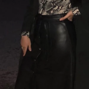 Svart kjol i skinnimitation - Snygg svart midikjol i skinnimitation med raka linjer och silverfärgade knappar framtill. Kjolen har hög midja, vilket ger en cool och edgy vibe.