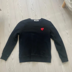 Svart sweatshirt Comme des Garçons Play - Svart sweatshirt från Comme des Garçons Play med det ikoniska röda hjärtat med ögon broderat på bröstet. Tröjan har rund hals, ribbade muddar och lång ärm. Tillverkad i mjuk bomull, perfekt för chill dagar. Snygg detalj med liten grön markering på ärmslutet.