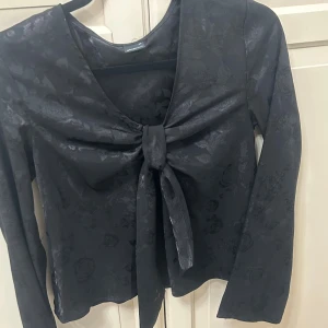 Svart blommig topp med knyt från Gina Tricot - Supersnygg svart topp från Gina Tricot med subtilt blommönster i tyget. Toppen har lång ärm, v-ringning och en stor knyt-detalj framtill som ger en trendig look. Perfekt att styla med jeans eller kjol för en cool outfit.(pris kan diskuteras vid snabb affär)