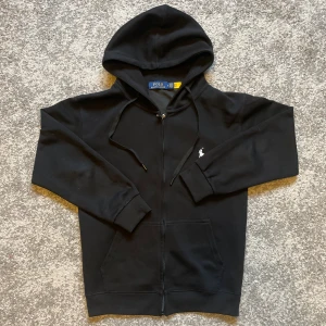 Svart hoodie från Polo Ralph Lauren - Svart hoodie från Polo Ralph Lauren med dragkedja, huva med snörning och klassisk vit broderad logga på bröstet. Tröjan har känguruficka framtill och är tillverkad i mjuk bomull. Perfekt för en chill och stilren look.