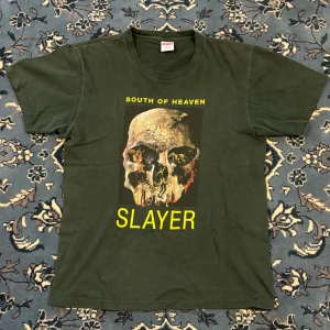Supreme x Slayer t-shirt mörkgrön M - Supreme t-shirt i mörkgrönt med Slayer South of Heaven tryck på framsidan och Supreme-logga på ryggen. Kortärmad, rund hals och tillverkad i 100% bomull. Grym grafik med dödskalle och detaljer i gult och rött, perfekt för dig som gillar streetwear och metal.