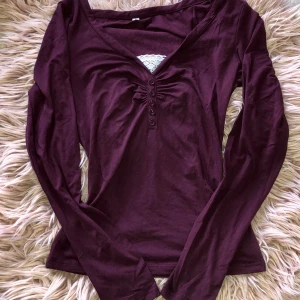 Vinröd långärmad topp med knappar - Vinröd långärmad topp med v-ringning och dekorativa knappar framtill. Toppen har en lätt rynkad detalj vid bysten och är tillverkad i mjukt bomullsmaterial. Perfekt för dig som gillar en enkel men snygg stil.