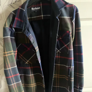 Rutig overshirt från Barbour i bomull - Snygg rutig overshirt från Barbour i 100% bomull. Jackan har mörkblå, gröna och röda toner med klassiskt rutmönster, två bröstfickor med knappar och mjuk känsla. Perfekt lager-på-lager-plagg för höst och vår.