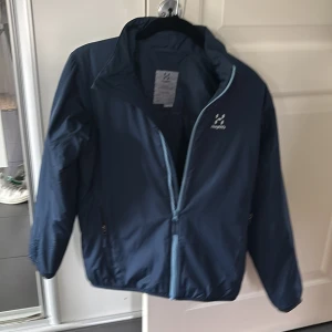 Blå vindjacka från Haglöfs - Snygg marinblå vindjacka från Haglöfs.Perfect nu inför höst. Jackan har en stilren design med diskret logga på bröstet och är tillverkad i lätt polyester. Perfekt för blåsiga dagar och enkel att slänga på sig över en hoodie. 