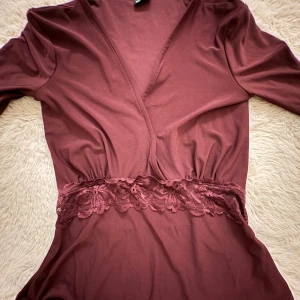 Vinröd omlottblus  med spet - Supersnygg vinröd omlottklänning från Gina Tricot i storlek XXS. blusen  har lång ärm och ett vackert spetsband i midjan som ger en extra touch. Materialet är mjukt och följsamt, perfekt för dig som gillar en feminin look med detaljer.