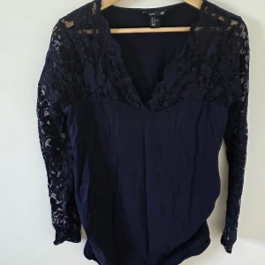 Svart spetsblus från H&M MAMA - Svart blus från H&M MAMA med långa ärmar i genombruten spets och v-ringad hals. Toppen har blommigt spetsmönster upptill och på ärmarna, medan nederdelen är i mjuk bomull. Perfekt för dig som vill ha en snygg och bekväm look.