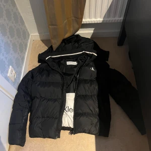 Svart pufferjacka från Calvin Klein - Säljer en svart pufferjacka från Calvin Klein med huva och dragkedja. Jackan har diskret CK-logga på bröstet och är vadderad för extra värme. Perfekt för kyliga dagar och har en stilren look som funkar till det mesta.