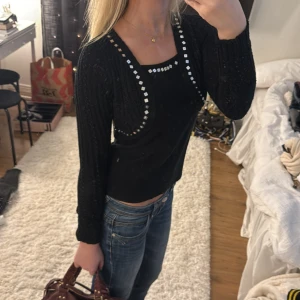 Svart bolerotopp med paljettdetaljer - Säljer en svart bolerotopp med långa ärmar och glittriga paljettdetaljer längs kanterna och halsringningen. Toppen är stickad och har en croppad passform som ger en snygg siluett.