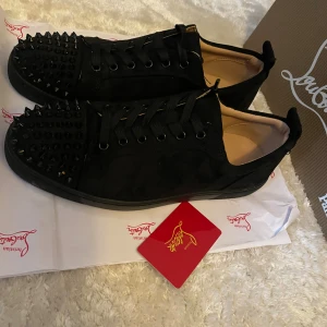 Christian Louboutin svarta sneakers med nitar - Svarta sneakers från Christian Louboutin i mocka med coola svarta nitar på tån och klassisk röd sula. Skorna har snörning och rund tå, och den lyxiga känslan förstärks av märkets ikoniska detaljer. Perfekta för dig som vill sticka ut med en edgy stil.