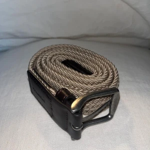 Beige flätat bälte från Pull&Bear - Snyggt beige flätat bälte från Pull&Bear med mörkbruna skinn-detaljer och metallspänne. Bältet har en stilren och modern look med twistad struktur som ger extra karaktär. Perfekt accessoar för att lyfta din outfit.