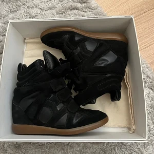Isabel Marant svarta sneakers med kilklack - Svarta sneakers från Isabel Marant i mocka och skinn med cool design och kilklack. Skorna har flera kardborreband, rund tå och beige sula – perfekta för dig som vill ha en trendig och sportig look med lite extra höjd. Köpta från NK Stockholm, tyvärr hittar jag inte det digitala kvittot längre. Skicket är 8/10, använda några månader under vintern 2024/2025. Jag säljer dem eftersom min syster, som de egentligen var tänkta till, har flyttat och de inte längre passar. Frakt skickas samma dag. 