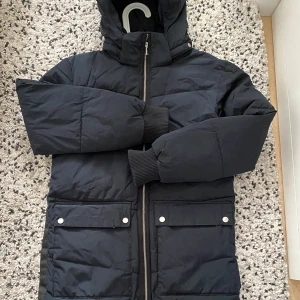 Svart lång pufferjacka från Everest - Svart lång pufferjacka från Everest med huva, två stora fickor med lock och tryckknappar samt dragkedja framtill. Jackan har ribbade muddar vid ärmsluten och en stilren, enkel design som håller dig varm under kalla dagar.