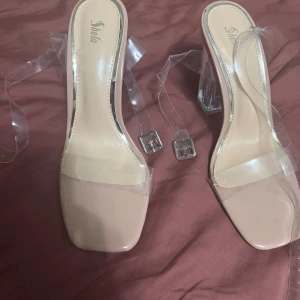 Beiga klackskor med transparent rem - Säljer ett par trendiga beiga klackskor från SheIn med transparenta remmar och fyrkantig tå. Skorna har en hög, genomskinlig blockklack och spänne i silver. Perfekta för dig som vill sticka ut med en modern och stilren look.