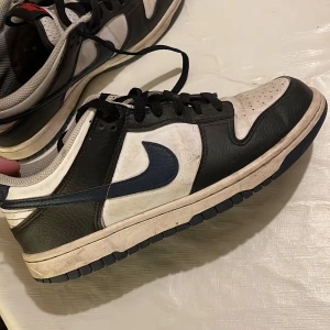 Nike Dunk Low svart/vit sneakers - Nike Dunk Low sneakers i svart och vitt med mörkblå swoosh och röda Nike-detaljer på plösen och hälen. Klassisk siluett med snörning, rund tå och platt sula. Ovandel i skinn och textil för en clean och tidlös look. Perfekta för dig som gillar streetstyle.