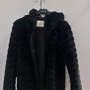 Svart pälsjacka med huva  - Mysig svart pälsjacka med huva och dragkedja. Jackan har ett fluffigt, randigt strukturmönster och är perfekt för kalla dagar. Snygg och trendig modell som håller dig varm hela vintern.