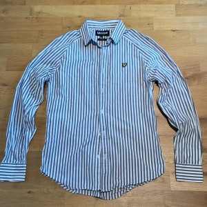 Randig skjorta från Lyle & Scott - Snygg slim fit skjorta från Lyle & Scott i vitt och grått med vertikala ränder. Klassisk krage, långa ärmar och knappar framtill. Ikonisk gul logga på bröstet. Perfekt för dig som gillar stilrena och fräscha plagg med lite edge. Storlek: S