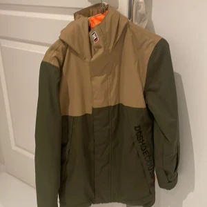 Grön och beige skid jacka från DC Shoes - Snygg grön och beige jacka från DC Shoes med huva och dragkedja. Perfekt för utomhusaktiviteter med sin stilrena design och praktiska funktioner så som att åka skidor .