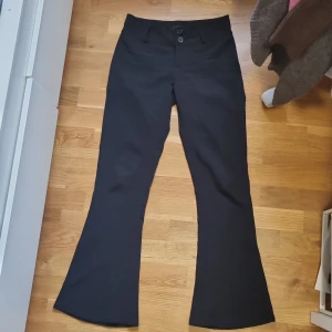 Svarta bootcut-byxor Gina Tricot S - Svarta bootcut-byxor från Gina Tricot i storlek S. Byxorna har hög midja, två knappar framtill, bälteshällor och fickor med lock bak. Snyggt utsvängda ben och klassisk look, perfekta till en trendig outfit.