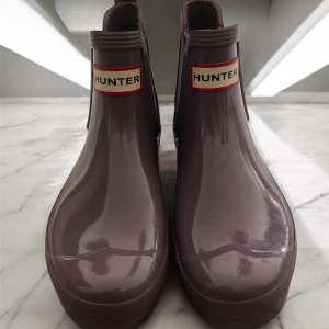 Lila Hunter chelsea boots i gummi - Snygga lila chelsea boots från Hunter i glansigt gummi. Klassisk design med resår på sidorna och tydlig Hunter-logga framtill. Perfekta för regniga dagar och har en platt sula för skön komfort.