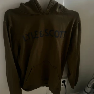  hoodie från Lyle & Scott - Säljer en olivgrön hoodie från Lyle & Scott Junior med stor logga i mörkblått framtill. Tröjan har huva, känguruficka och långa ärmar. Materialet är mjuk bomull och passformen är normal. Perfekt för chill dagar och streetwear.