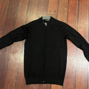 Svart kofta med dragkedja Keepman - Svart kofta köpt i i Italien, från Keepman med hel dragkedja framtill och ribbad grå krage. Stilren och enkel design som funkar till många outfits. Långärmad och tillverkad i mjukt material som känns skönt mot huden.