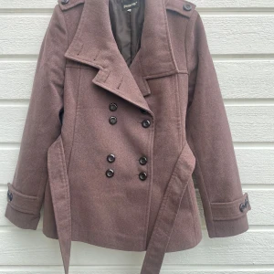 Brun kortare höstkappa  - Stilren brun kortare höstkappa från Piero med dubbelknäppning, axelklaffar och bälte i midjan. Jackan har långa ärmar, dekorativa knappar vid ärmslut och är tillverkad i en mjuk blandning av viskos, polyester och polyamid. Liten i storleken. Mer lik storlek S 