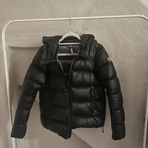 Svart Colmar pufferjacka med huva - Säljer en svart pufferjacka från Colmar Originals med huva och dragkedja. Jackan har en glansig finish, breda quiltade sektioner och logga på ärmen. Perfekt för kalla dagar och riktigt snygg till streetwear-stilen.
