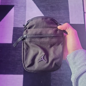 Svart axelväska från Jordan - Svart axelväska från Jordan med klassisk Jumpman-logga på framsidan. Väskan har en kompakt, rektangulär form och är tillverkad i slitstark polyester. Utrustad med dragkedja och justerbar axelrem, perfekt för att bära dina essentials.