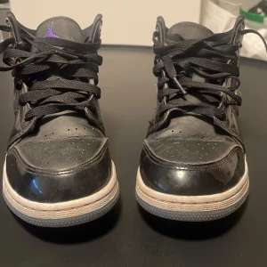 Nike Air Jordan 1 Mid Svart/Lila - Säljer ett par Nike Air Jordan 1 Mid i svart med lila detaljer. Skorna har klassisk Jordan-logga på sidan, svart snörning och en vit/grå sula med genomskinlig undersida. Ovandelen är i blankt skinn och textil, vilket ger en clean och sportig vibe.