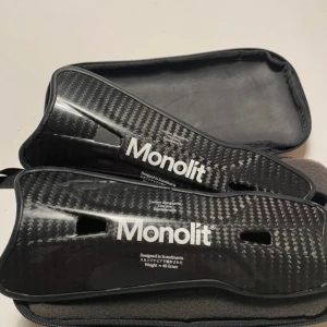 Monolit benskydd i kolfiber - Säljer ett par svarta benskydd från Monolit i kolfiber med snyggt mönster och vit logga. De är lätta, har en ergonomisk form och kommer i ett smidigt fodral. Perfekta för dig som vill ha riktigt schyssta skydd på planen.