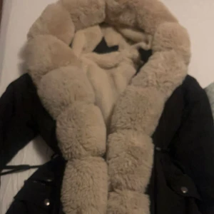 Svart parkas med beige fuskpäls - Säljer en svart parkas med stor, fluffig beige fuskpäls både på utsidan och  insida. Jackan har dragsko i midjan, fickor med knappar och en riktigt mysig vibe. Perfekt för kalla dagar när du vill vara både varm och snygg.