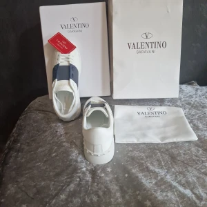 Valentino Garavani vita sneakers - Snygga vita Valentino Garavani sneakers med bred mörkblågrå rem över ovansidan och ikoniska nitar på hälen. Skorna har rund tå, platt sula och är tillverkade i lyxigt skinn. Perfekta för dig som vill ha en clean och exklusiv look.ingen låda.