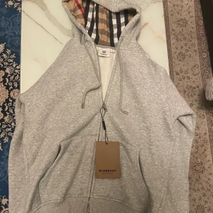 Grå Burberry hoodie med rutig huva - Säljer en grå hoodie från Burberry i storlek S. Tröjan har dragkedja, snörning vid huvan och den klassiska Burberry-rutiga insidan på huvan. Materialet är mjukt och känns lyxigt, perfekt för dig som vill ha något stilrent med en twist.
