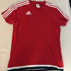 Röd Adidas t-shirt M - Snygg röd fotbollströja från Adidas i storlek M. Klassisk design med vita ränder på axlarna och Adidas-logga på bröstet. Tröjan har rund hals, korta ärmar och vita samt svarta detaljer längst ner. Tillverkad i lätt och ventilerande material.