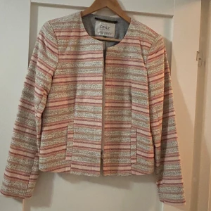 Randig kavajjacka från ONLY, strl 38 - Snygg kavajjacka från ONLY i storlek 38. Jackan har ett randigt mönster i rosa, beige och grå toner, rund halsringning och långa ärmar. Den är tillverkad i ett vävt material och har en stilren, öppen front utan knappar.