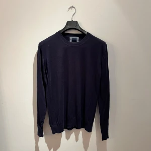 Mörkblå stickad tröja från Redgreen - En stilren mörkblå stickad tröja från Redgreen i klassisk modell. Tröjan har rund halsringning, ribbade muddar och lång ärm. Perfekt för lager-på-lager och enkel att matcha med jeans eller chinos. Hör av er vid frågor